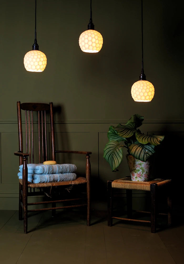 Triple cluster of beehive style porcelain bathroom pendant lights casting shadows with wooden chair, bath towels and plant.