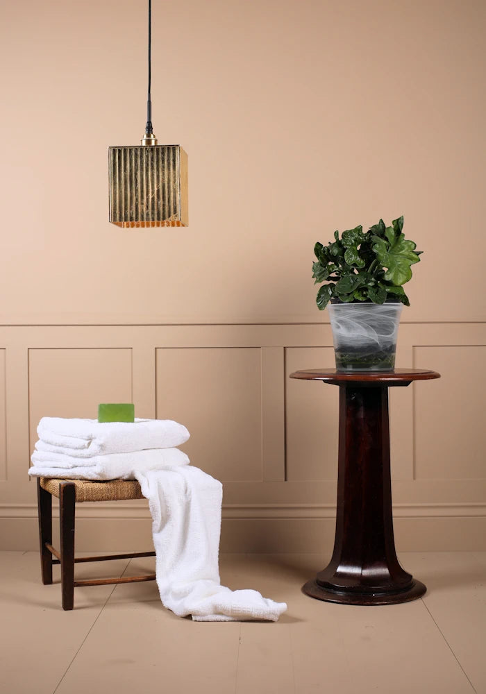 Large cube gold leaf bathroom pendant light switched off with bath towels and plant on a stand.