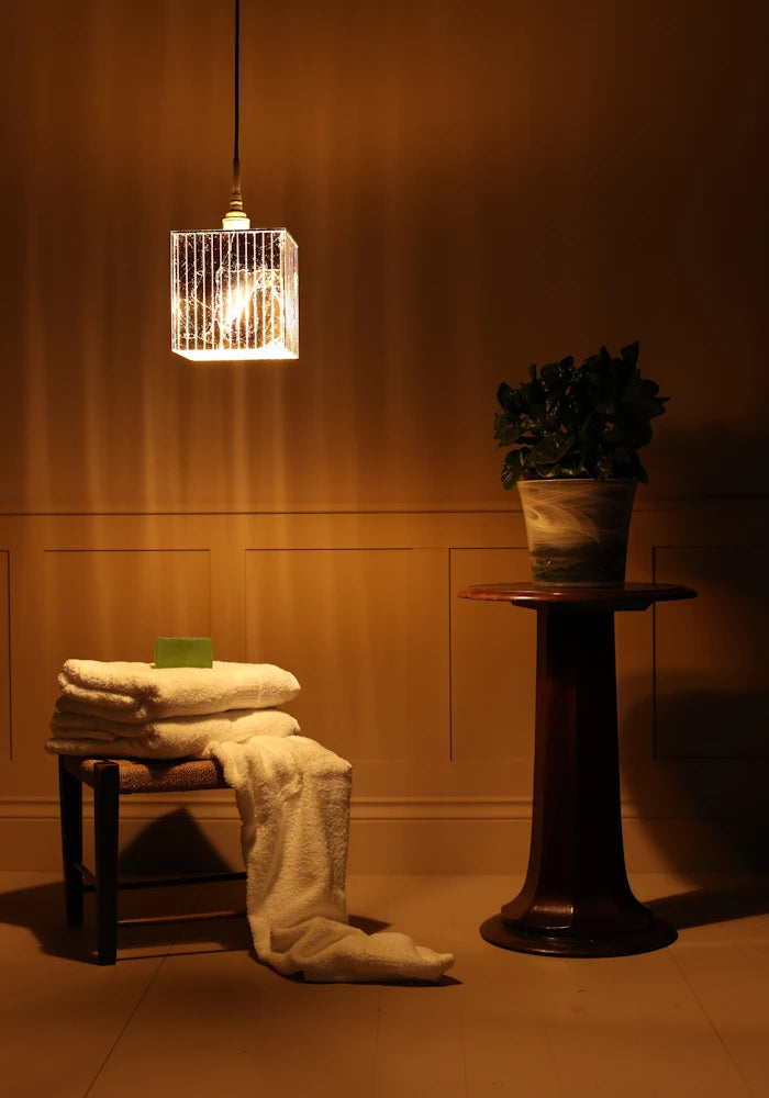 Large cube gold leaf bathroom pendant light casting shadows onto bath towels and plant on a stand.