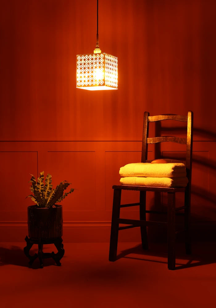 Large square Moroccan patterned gold bathroom pendant light casting shadows, plant and wooden chair with towels.