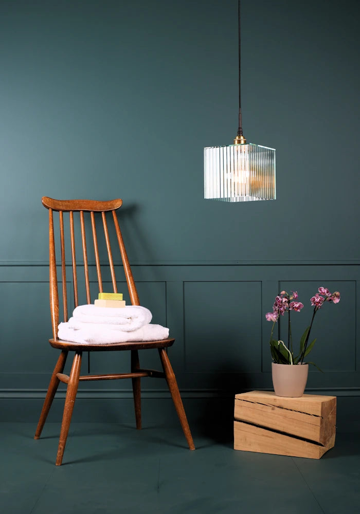 Square ribbed glass bathroom pendant light switched off with wooden chair, bath towels and plant.