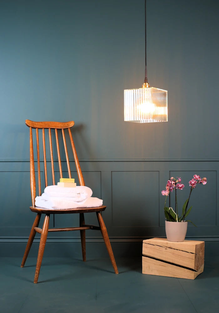 Square ribbed glass bathroom pendant light with wooden chair, bath towels and plant.