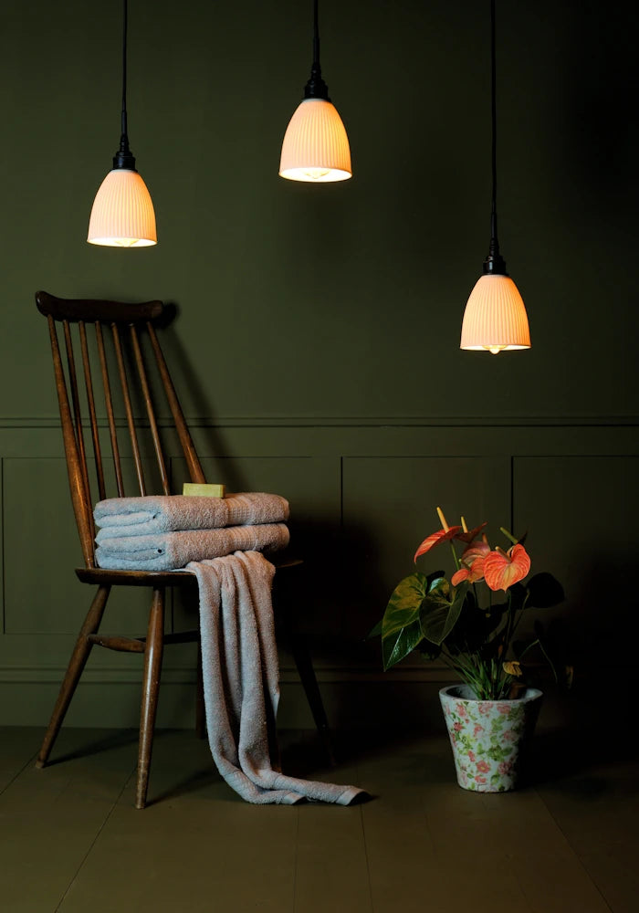 Triple porcelain cluster bathroom pendant light casting shadows onto wooden chair, bath towels and plant.