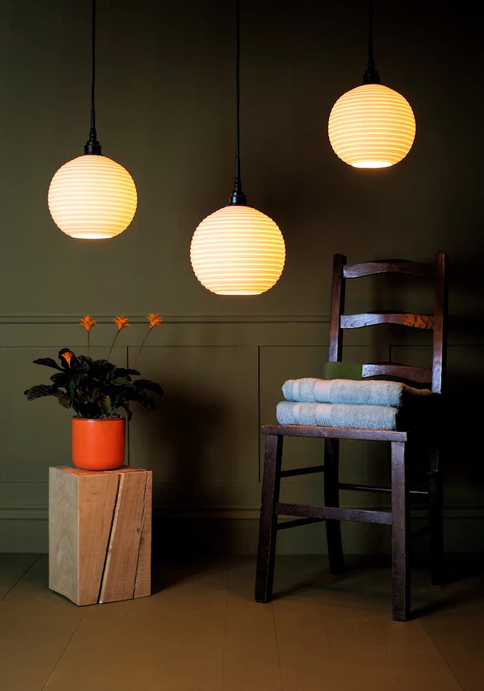 Large triple spherical porcelain bathroom pendant light cluster casting shadows onto plant, wooden chair and bath towels.