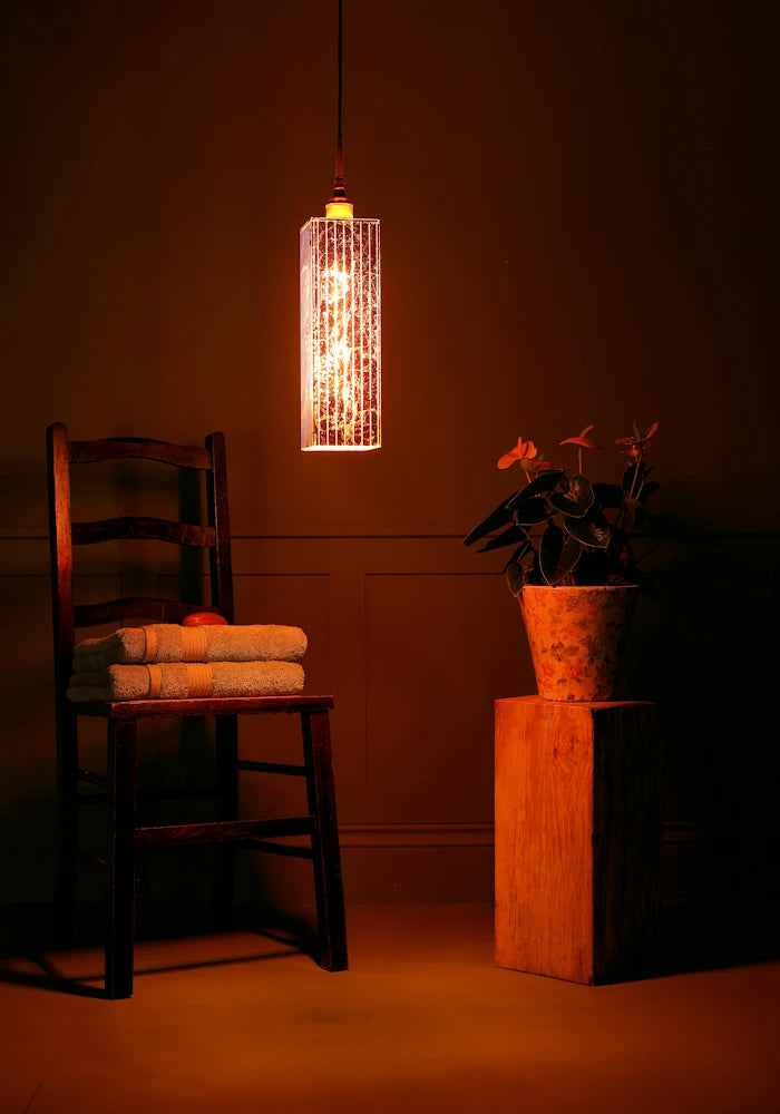 Elongated rectangular copper leaf bathroom pendant light, wooden chair, towels and plant.