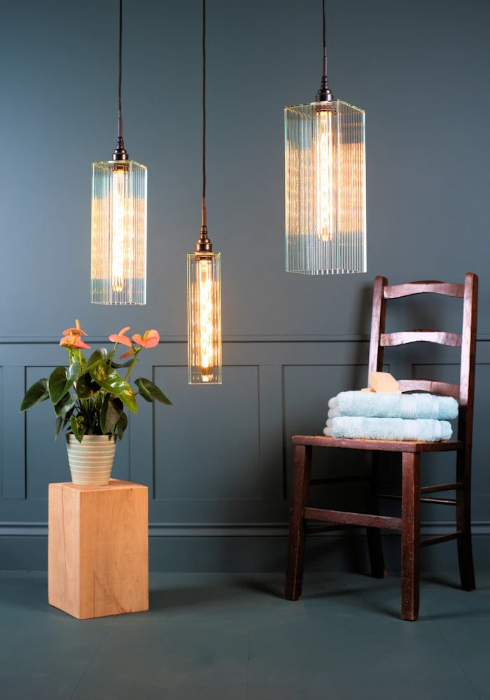 Triple large ribbed glass bathroom pendant light cluster with plant, wooden chair and towels.