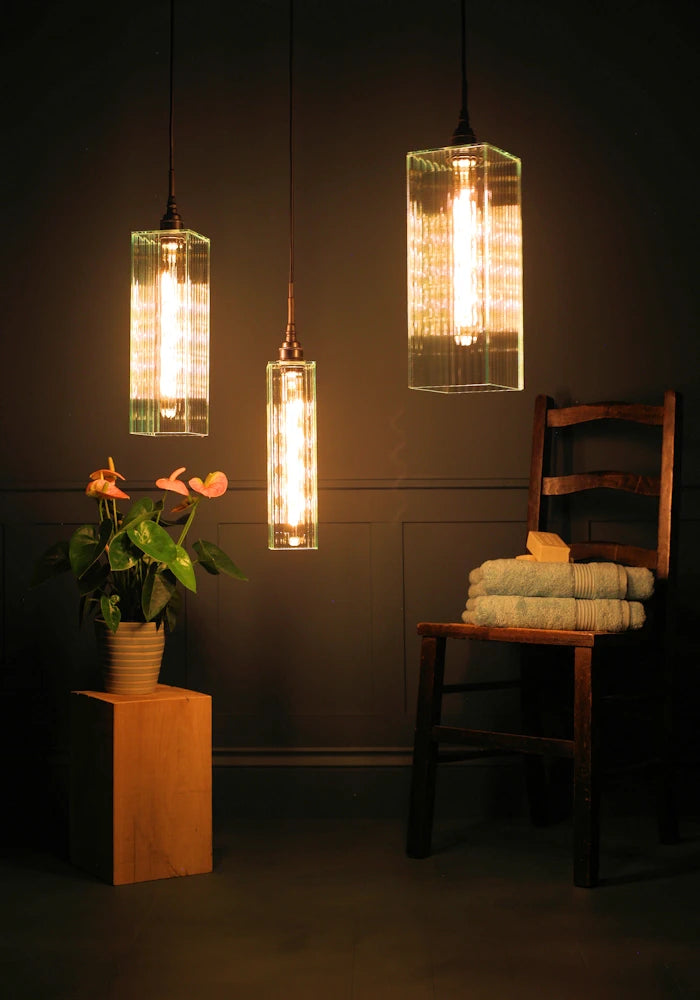 Triple large ribbed glass bathroom pendant light cluster casting shadows onto plant, wooden chair and towels.