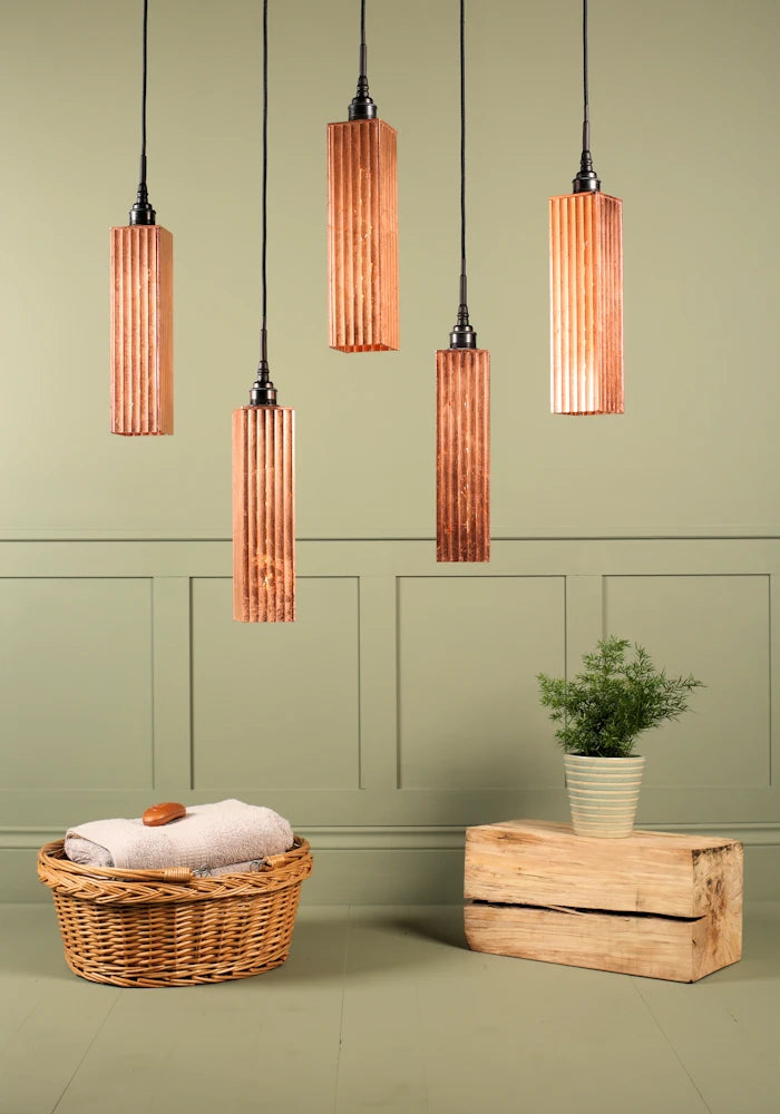 Cluster of five long rectangular copper leaf bathroom pendant lights switched off, basket of bath towels and plant.