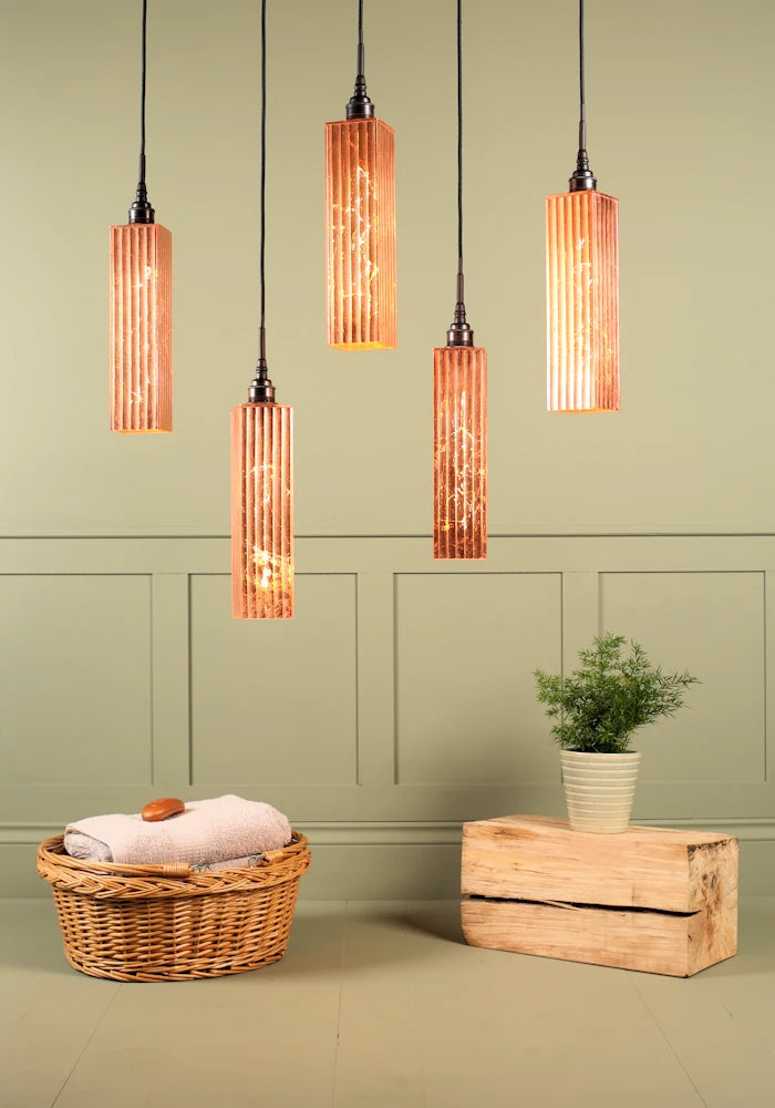 Cluster of five long rectangular copper leaf bathroom pendant lights, basket of bath towels and plant.