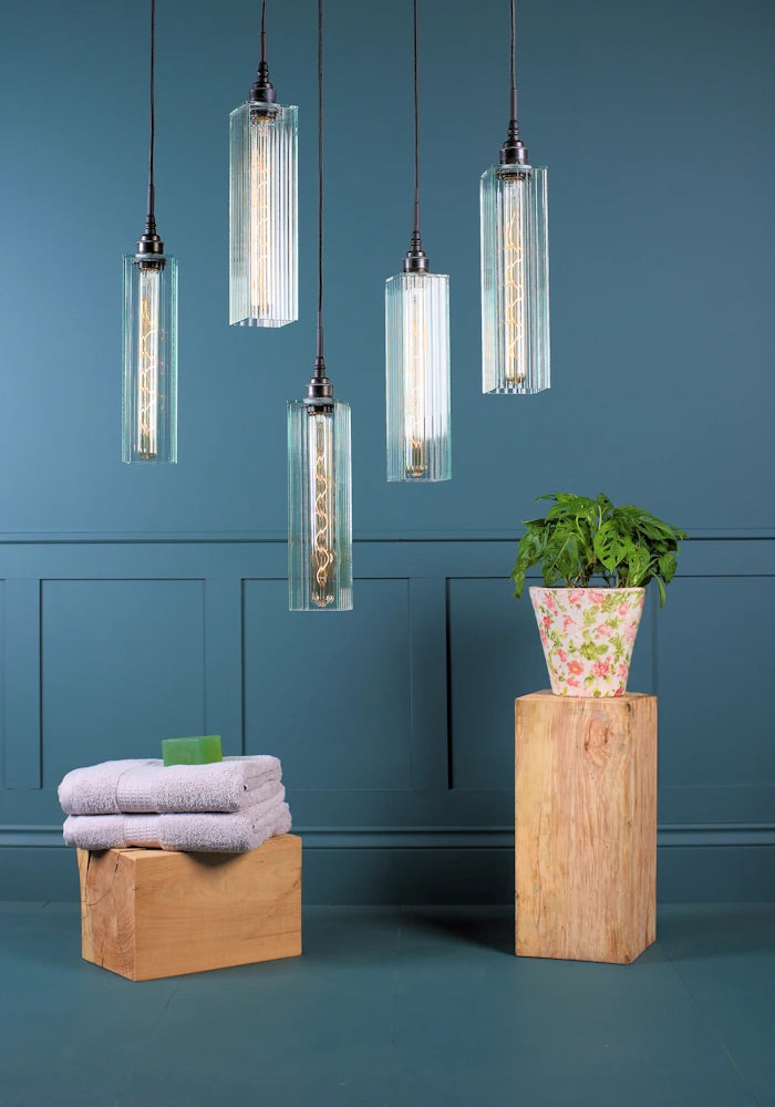 Five long rectangular ribbed glass cluster bathroom pendant lights switched off with bath towels and pot plant.