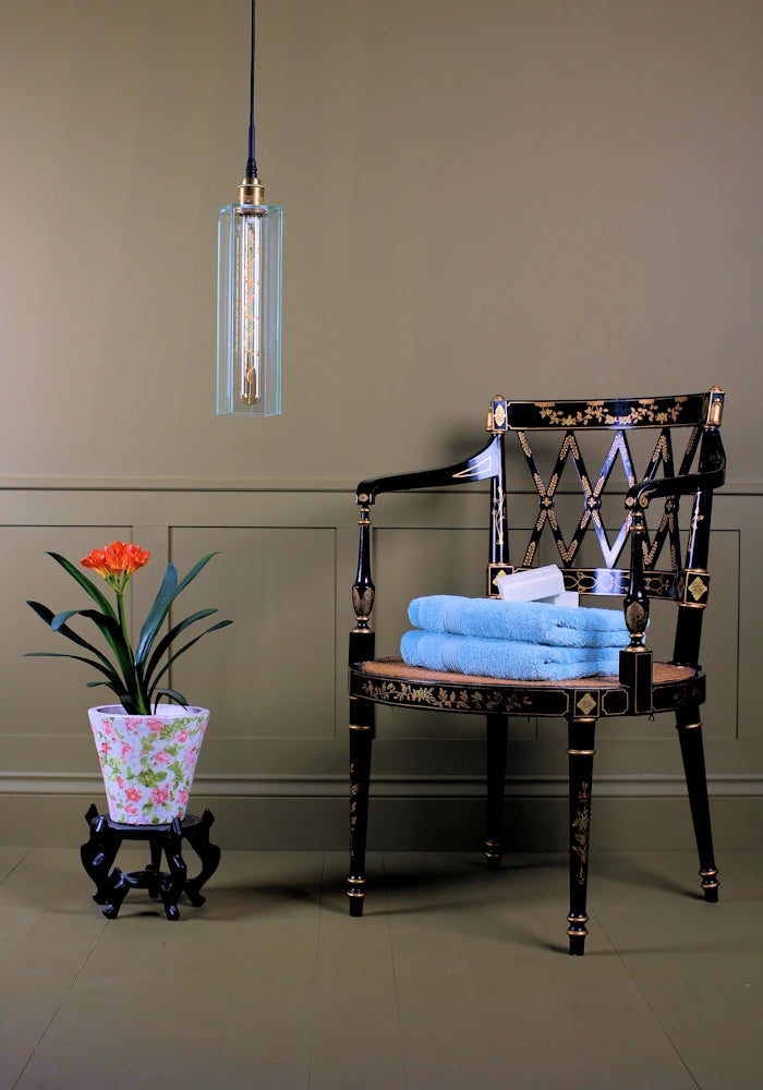 Long clear glass bathroom pendant light switched of with tropical plant, ornate chair and bath towels.