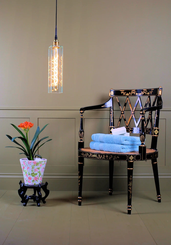 Long clear glass bathroom pendant light with tropical plant, ornate chair and bath towels.