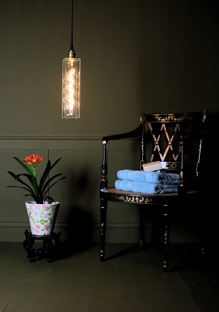 Long clear glass bathroom pendant light casting shadows on tropical plant, ornate chair and bath towels.