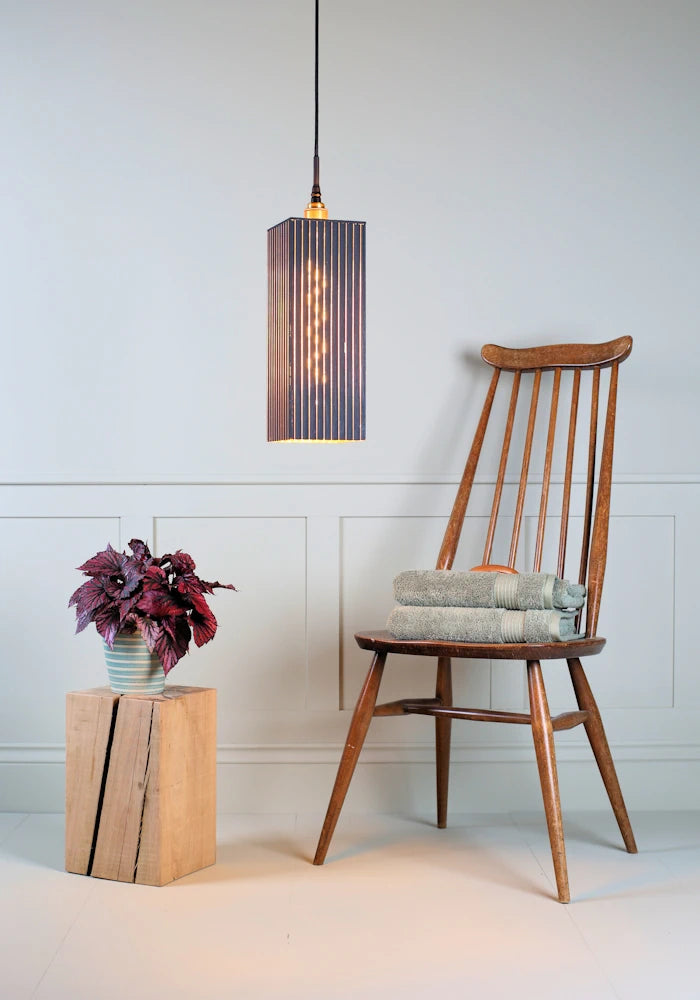 Large black and gold rectangular bathroom pendant light with plant, wooden chair and bath towels.