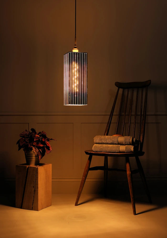 Large black and gold rectangular bathroom pendant light casting shadows onto plant, wooden chair and bath towels.