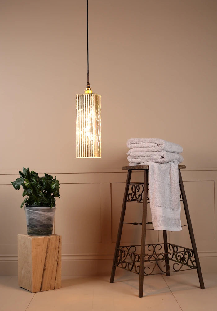 Long rectangular gold leaf bathroom pendant light casting with bath towels and plant.