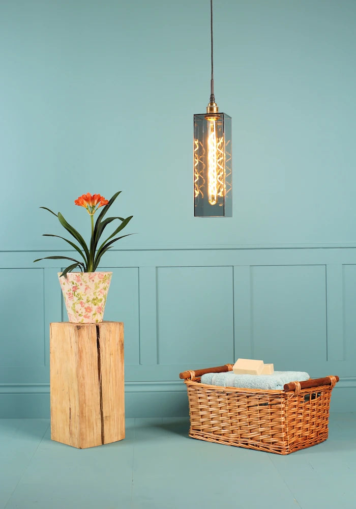 Elongated rectangular smoked glass bathroom pendant light, plant, wooden basket and bath towels.