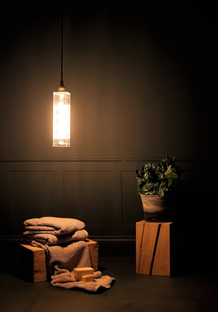 Long rectangular ribbed glass bathroom pendant light casting shadows onto bath towels and plant.