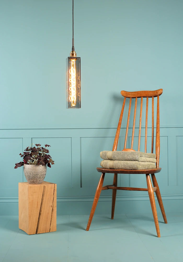 Elongated rectangular smoked glass bathroom pendant light, plant, wooden chair and bath towels.